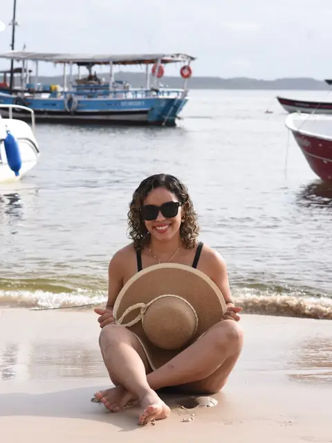 Viajante de chapéu sentada na praia em Morro de São Paulo, com barcos ao fundo